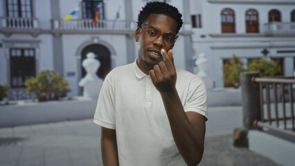 Man in white polo shirt wearing glasses making finger heart gesture in front of building entrance; affection.