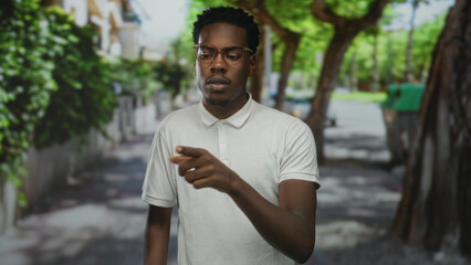 Fototapeta premium Young man wearing glasses and white polo shirt pointing finger to object on a sunlit city street; confidence.