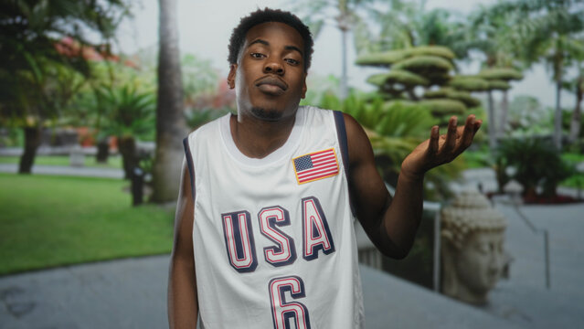 Man holds up bare hand in lush green park wearing white usa basketball jersey patched with american flag; confusion.