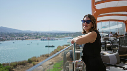 Obraz premium Woman resting bare arms on glass railing on cruise terrace overlooking marina and sea; relaxation travel.