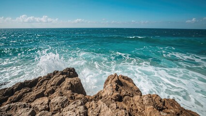 A view of the ocean from the rocky shore with waves crashing, under a partly cloudy sky.