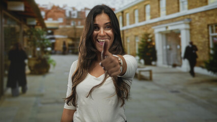 Fototapeta premium Woman giving thumbs up toward camera on street with urban shops and blurred passersby; confidence support.