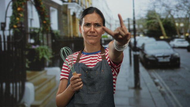 Woman in red and white striped shirt and denim apron holding a whisk and making rock horns on street with parked cars and iron railings; joy baking creativity.