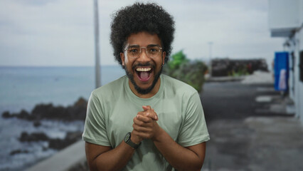 Smiling man on seaside promenade in casual attire enjoying beach view outdoors with cheerful expression and joyful demeanor against a scenic sea background under a cloudy sky.