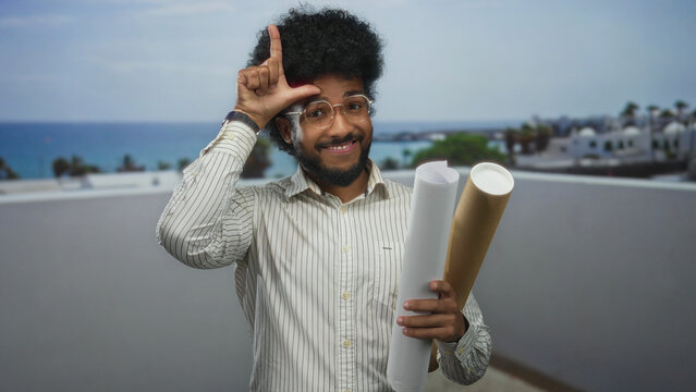 Smiling man with blueprints makes a loser sign at beach, combining architecture and humor in a serene seaside setting with a playful expression.