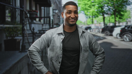 Young black man posing with hands on hips, wearing jacket and black tshirt on street; confidence approachability warmth.