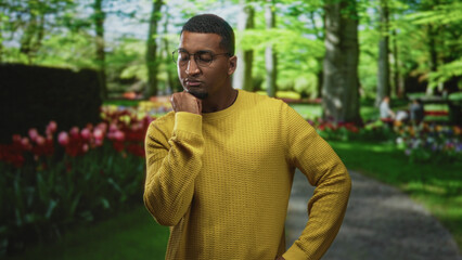Young black man hand on chin thinking in forest wearing yellow sweater and glasses; thoughtful reflection.