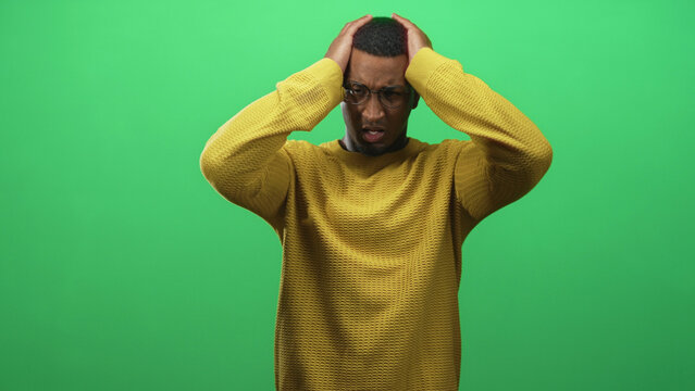Black man in yellow sweater clutching head with both hands in studio green screen, wearing glasses and a pained expression; stress tension.