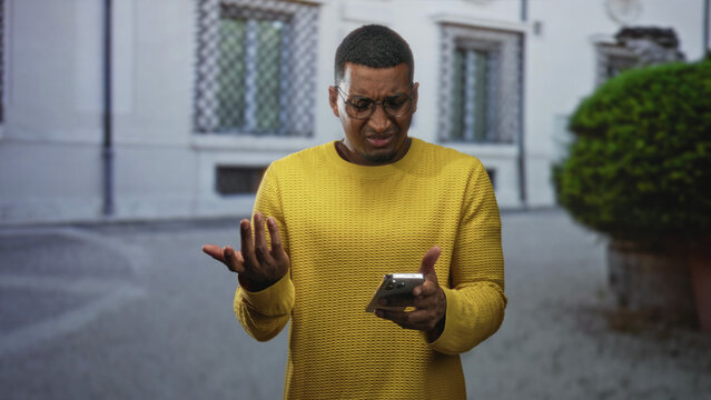 Young black man wearing yellow sweater and glasses holding smartphone with palm up questioning gesture on street in front of a building; frustration.