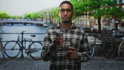 Young black man holding smartphone and coffee cup on street by canal in city, wearing glasses and plaid shirt; calm reflection.