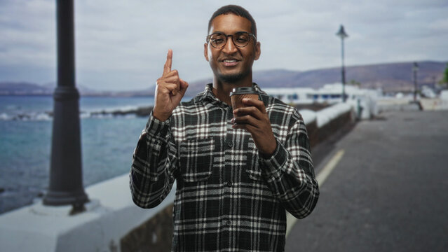 Man holding coffee cup in hand on a coastal street, pointing finger while standing and smiling; calm confidence.