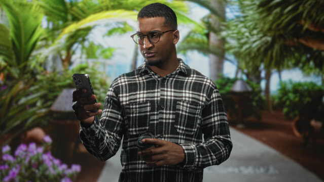 Young black man holds smartphone and coffee cup while looking at the screen in studio; thoughtful concentration.