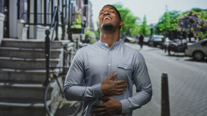 Fototapeta premium Young black man laughing with hand on chest and other hand on abdomen, wearing a button shirt by steps and bicycle on a street with parked cars; amusement.