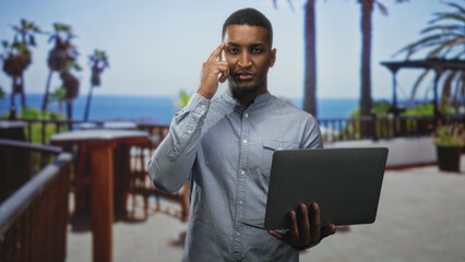 Young black man holding laptop and pointing finger to temple on a building balcony by the ocean with palm trees; focused remote work.