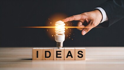 Hand reaching toward glowing light bulb above wooden blocks spelling “IDEAS” on dark background.