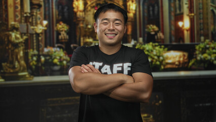 Young chinese man smiling and wearing staff tshirt crosses arms in church building; service faith devotion.