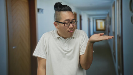 Man in white polo holding palm up presenting open hand in hotel building corridor, wearing glasses and bun; skepticism decision choice.