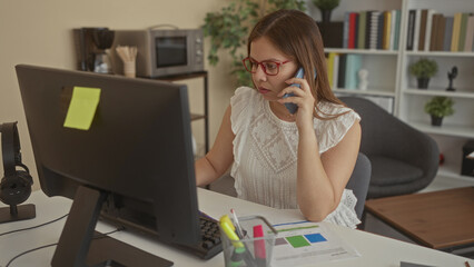 Woman holds phone to ear while reviewing printed charts and looking at computer monitor at office...