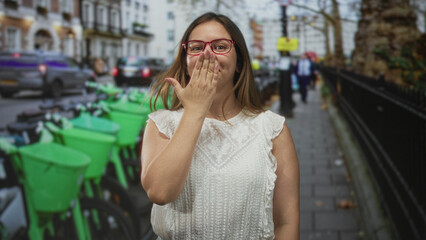 Fototapeta premium Woman in white blouse wearing red glasses covering mouth with hand among rows of green rental bikes parked on a city street; playful charm.