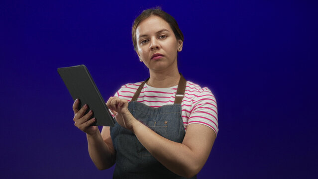 Young caucasian woman in denim apron and pink striped shirt tapping a tablet with fingers in a studio with deep blue backdrop; concentration.
