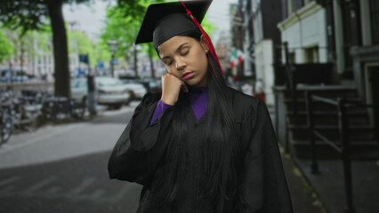 Young latin woman in graduation cap and gown touches chin while standing on city street;...