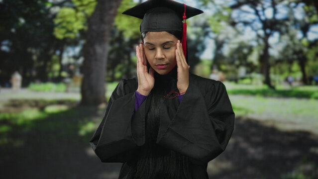 Young woman in black graduation gown and cap raises hand outdoors in lush green forest; pride accomplishment.