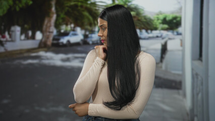 Latin woman touching chin in a thoughtful pose on city street with parked cars and long dark hair; uncertainty.
