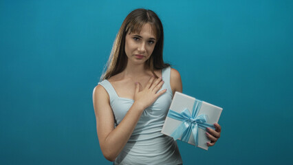 Young woman holding a gift box with blue ribbon, hand to chest posed in studio with teal backdrop; sincere gratitude.