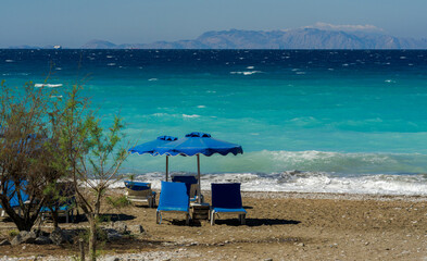 Sunny beach with blue sun loungers and umbrella facing turquoise sea waves, with distant mountains on the horizon and a small tree framing the seaside view.