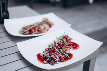 A gourmet plated dish featuring sliced meat topped with a vibrant red berry sauce and fresh microgreens, served on elegant white plates in a modern dining setting.