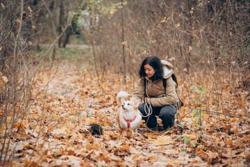 Woman with dark hair kneeling in a forest path covered with autumn leaves, interacting with a small dog, showcasing a joyful moment in nature during a leisurely walk