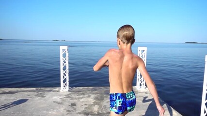 An excited young boy in blue swim trunks joyfully jumps into a calm lake on a sunny summer day, making a playful hand gesture. A carefree moment of childhood fun and vacation adventure.