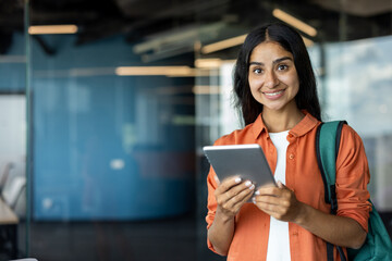 Portrait of a young Indian female student standing with a backpack indoors, holding a tablet, smiling at the camera