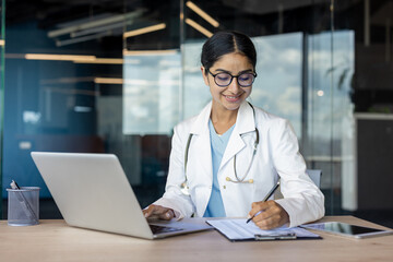 Indian young female doctor sitting in hospital office at laptop and taking notes in notebook