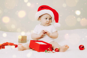 Baby wearing a red Santa hat is surrounded by Christmas gift boxes and decorations, seated on a white bed. The child shows curiosity and delight in the festive setting.