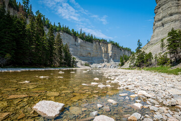 Naklejka premium Steep Canyon, Stone Cliffs, Shallow River, Fallen Boulders, and Boreal Forest Under a Blue Sky near to Vaureal Waterfall on a Sunny Day in Anticosti, Quebec, Canda