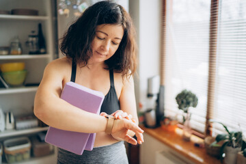 Athletic woman in black sportswear checking fitness tracker while holding yoga block in a bright room with natural light, promoting wellness and healthy lifestyle choices