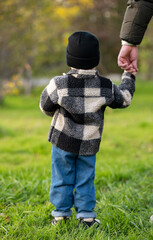 Young child holding an adult’s hand while standing on a grassy field. Dressed warmly, the child gazes ahead, evoking themes of care, safety, and connection in a natural outdoor setting.