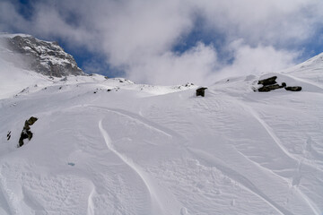 Snow-covered alpine slope with scattered rocks, strong wind patterns, and fog moving around a rugged mountain. Atmospheric winter landscape in cold high-altitude conditions.