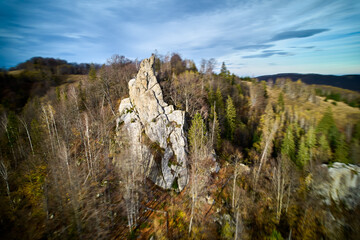 Dynamic aerial shot of towering, jagged rock formation amid mix of bare and evergreen trees. Dramatic rock spire contrasts with surrounding forest and rolling hills under vast, cloudy sky.