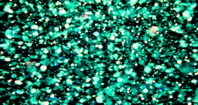 Particles float in water over a dark background, reflecting top light in green and blue hues, creating a glittering, halo-like, cosmic universe effect.
