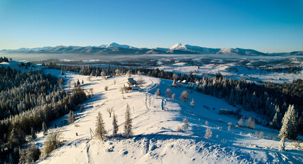 Aerial view reveals stunning winter landscape with snow-covered hills and dense forests. Houses nestled among trees, while background features majestic range of mountains under clear blue sky.