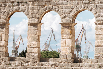 Ancient stone arches framing industrial port cranes on a sunny day, contrasting historic architecture with modern maritime infrastructure.