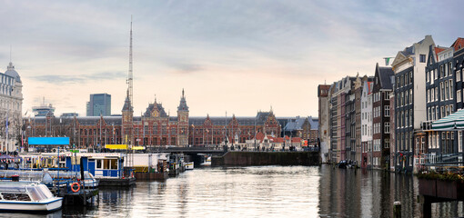 Twilight at Amsterdam Central Station's harbor, with canal boats gliding on water, historic architecture, and reflections sparkling under the evening lights.