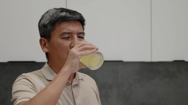 Asian man drinking fresh lemon juice from clear glass in modern kitchen. Ideal for wellness content, morning routines, natural drinks, detox themes, and healthy lifestyle materials.