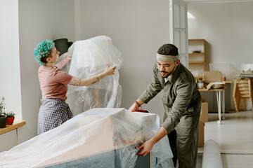 Young adult Caucasian woman and young adult man unpacking furniture and covering items with plastic sheet while moving and renovating apartment, working together in modern home