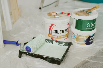 Closeup of paint roller and buckets with green paint resting on plastic sheet, showing preparation for home renovation by young couple moving into new living space © AnnaStills