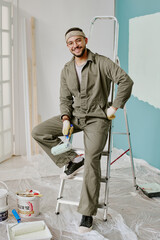 Portrait of young adult man smiling while sitting on ladder holding paint roller, wearing work...