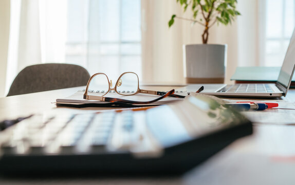 Eyeglasses and calculator close-up and documents with modern laptop sitting at desk background next bright daylight window. Business, home finances money savings