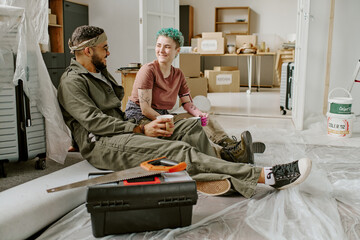 Woman with short blue hair and young adult man sitting on floor drinking coffee and smiling while taking break from renovations in new home